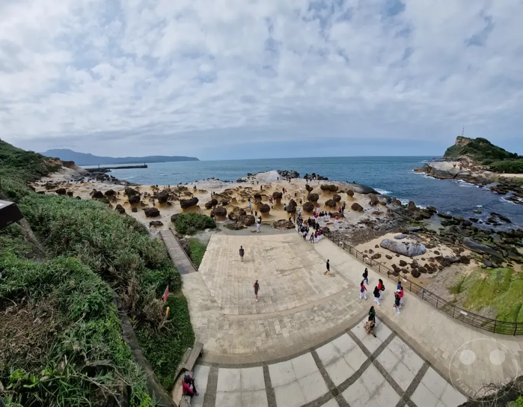 Die einzigartige Küstenlandschaft des Yehliu Geoparks in Taiwan präsentiert sich mit ihren charakteristischen Pilzkopf-Felsen und einer belebten Atmosphäre, während Besucher die skurrilen Formationen erkunden. Das weite Meer und der bewölkte Himmel erzeugen eine dramatische Kulisse für die geologischen Wunder. Die Szene strahlt die Faszination eines beliebten Naturdenkmals aus, das von Menschen aus aller Welt besucht wird.