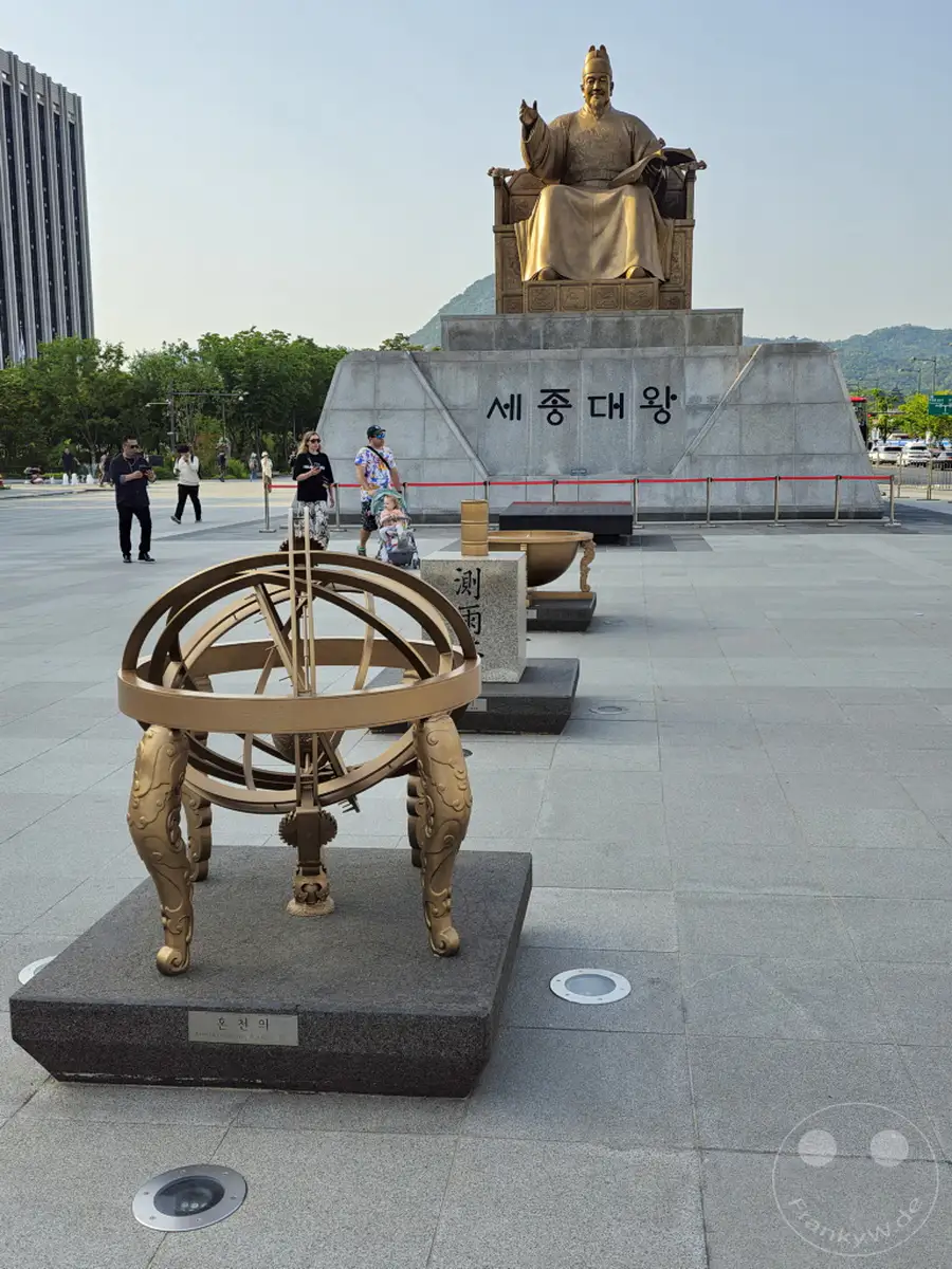 In Seoul, Südkorea, steht das imposante Denkmal für König Sejong den Großen auf dem Gwanghwamun Square. Vor dem Denkmal sind detailreiche astronomische Instrumente ausgestellt, die an die wissenschaftlichen Errungenschaften der Joseon-Dynastie erinnern. Die Szene wird von Besuchern belebt, die die historische Stätte erkunden.