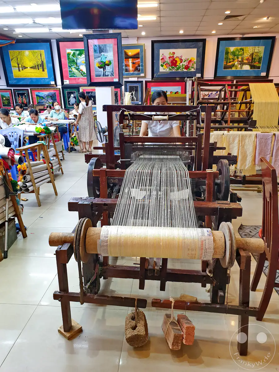 Ein traditioneller Seidenwebstuhl wird in einer Werkstatt in Dai Bat, Vietnam, verwendet, um feine Stoffe herzustellen. Mehrere Frauen sind an der Herstellung beteiligt, was die gemeinschaftliche und handwerkliche Natur der Seidenproduktion unterstreicht. Die Szene strahlt eine Atmosphäre konzentrierter Arbeit und kultureller Bewahrung aus.