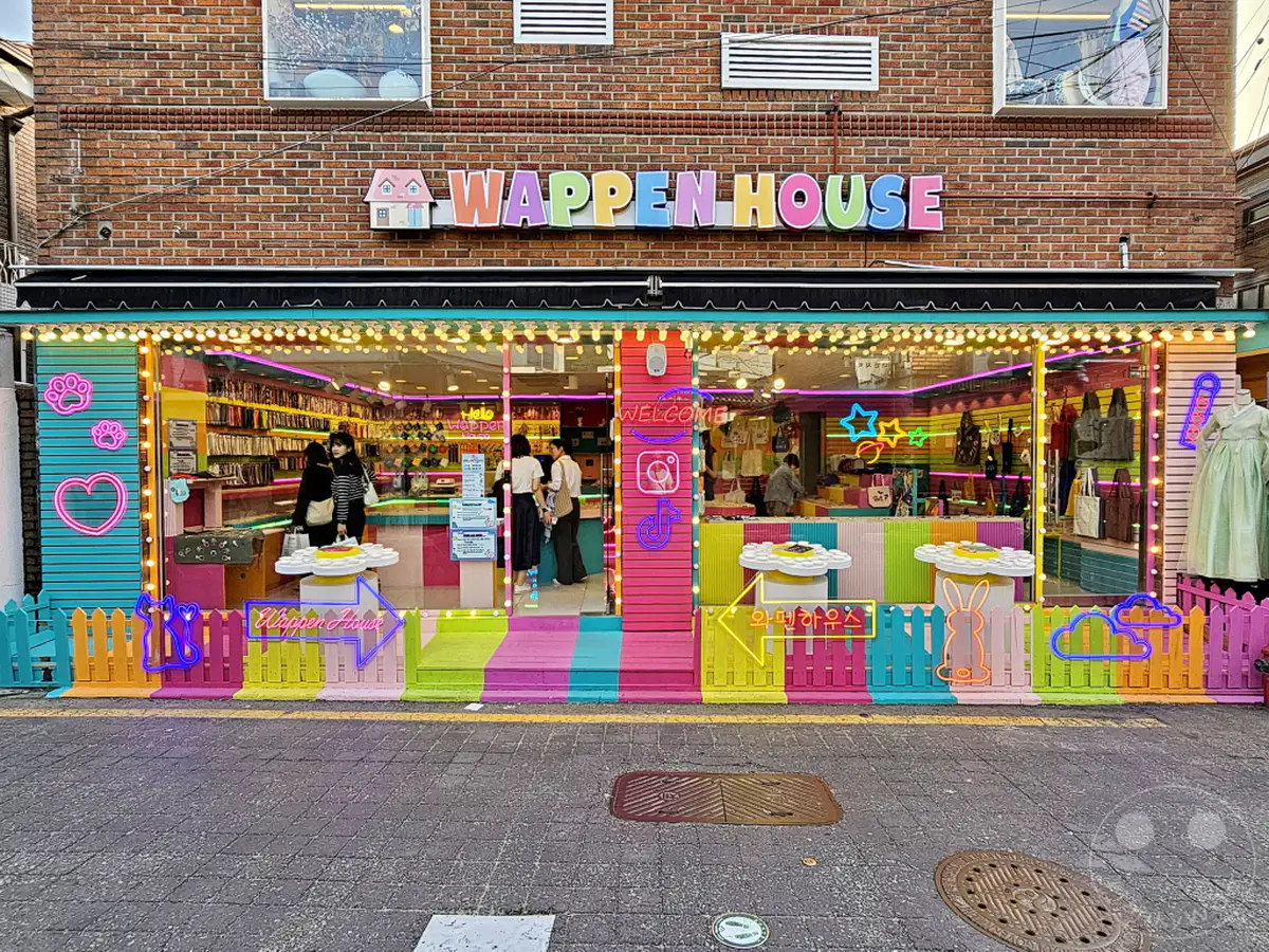 Die lebendige und verspielte Fassade eines "Wappen House" Shops in Südkorea, Seoul, Bukchon Hanok Village, sticht durch seine leuchtenden Neonlichter und eine pastellfarbene Dekoration hervor. Eingelassene Regale voller Produkte sind durch die Glasfronten sichtbar, während eine bunte Gartenzwerg-ähnliche Zäunung den Eingangsbereich säumt. Die fröhliche und einladende Atmosphäre des Geschäfts wird durch die symmetrische Anordnung und die künstlerische Gestaltung verstärkt.