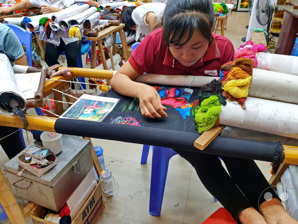 In Vietnam, Dai Bat, widmet sich eine Handwerkerin der feinen Kunst der Seidenstickerei. Bunte Garnknäuel liegen bereit, um auf den Stoff gebracht zu werden, während eine Vorlage das zu stickende Motiv vorgibt. Die konzentrierte Atmosphäre unterstreicht die Sorgfalt und das Engagement, die in diesem traditionellen Handwerk stecken.