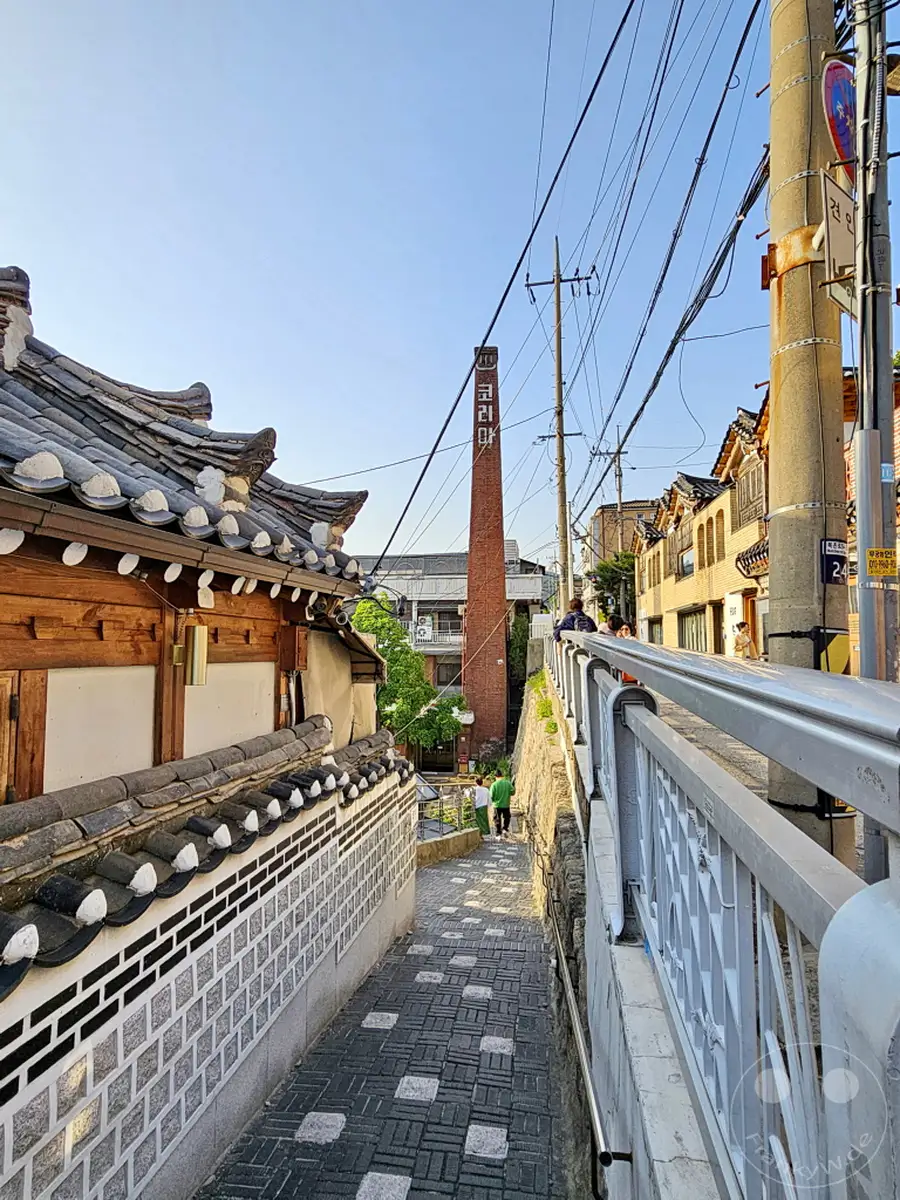 Eine schmale, gepflasterte Gasse windet sich bergab durch das traditionelle Bukchon Hanok Village in Seoul, Südkorea. An den Seiten reihen sich kunstvoll gedeckte Dächer von Hanok-Häusern und moderne Gebäude, während ein hoher, roter Backsteinschornstein mit koreanischer Schrift den Himmel durchbricht. Die Szene strahlt eine ruhige und authentische Atmosphäre aus, die die Verschmelzung von Geschichte und modernem Leben in der koreanischen Hauptstadt widerspiegelt.