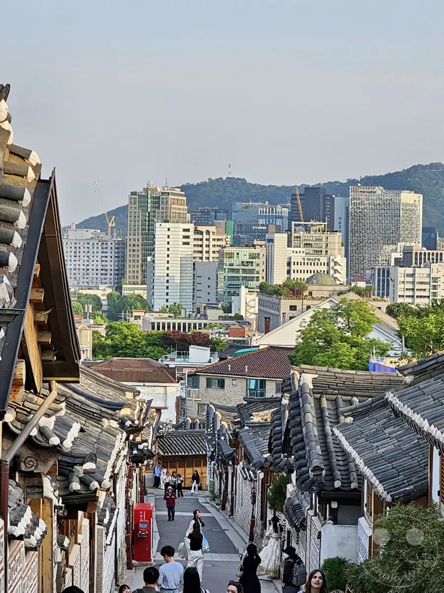 Die Aufnahme gibt einen malerischen Blick auf eine Gasse im Bukchon Hanok Village in Seoul, Südkorea frei, gesäumt von traditionellen koreanischen Häusern mit ihren charakteristischen Dächern. Im Hintergrund erheben sich moderne Hochhäuser und bilden einen faszinierenden Kontrast zur historischen Umgebung. Die Szene fängt eine harmonische Verschmelzung von Altem und Neuem in der pulsierenden Metropole ein, während Menschen die Gasse entlangschlendern und das Flair genießen.