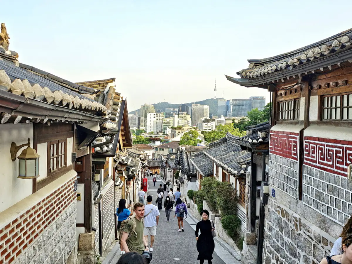 Ein malerischer Blick auf die engen Gassen des Bukchon Hanok Village in Seoul, Südkorea, mit traditionellen koreanischen Häusern (Hanoks), die von modernen Hochhäusern im Hintergrund eingerahmt werden. Touristen schlendern durch die historische Umgebung und genießen die einzigartige Mischung aus Alt und Neu. Die Szene strahlt eine friedliche und kulturell reiche Atmosphäre aus, die Besucher aus aller Welt anzieht.