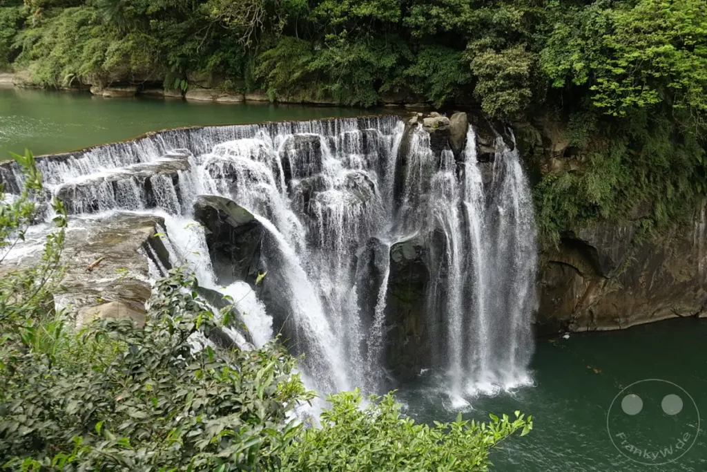 Der Shifen Wasserfall in Taiwan stürzt breit und kraftvoll über mehrere Ebenen in ein smaragdgrünes Becken. Umgeben von üppiger Vegetation und felsigen Hängen entfaltet sich eine majestätische Naturkulisse. Die Szenerie strahlt eine friedliche und erfrischende Atmosphäre aus, die die Schönheit der Landschaft unterstreicht.