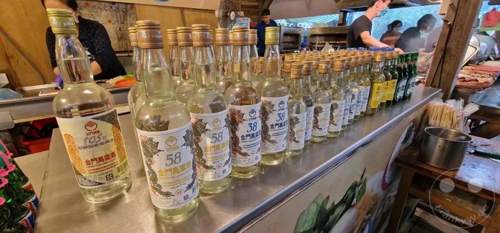 Eine Reihe von Flaschen mit Kaoliang-Schnaps steht in einem Geschäft in Jiufen Old Street, Taiwan, zum Verkauf. Die Flaschen sind mit Drachenmotiven und der Alkoholstärke verziert, was auf eine traditionelle Herstellung hindeutet. Die Szene vermittelt die geschäftige Atmosphäre eines lokalen Marktes, auf dem Touristen und Einheimische einkaufen.