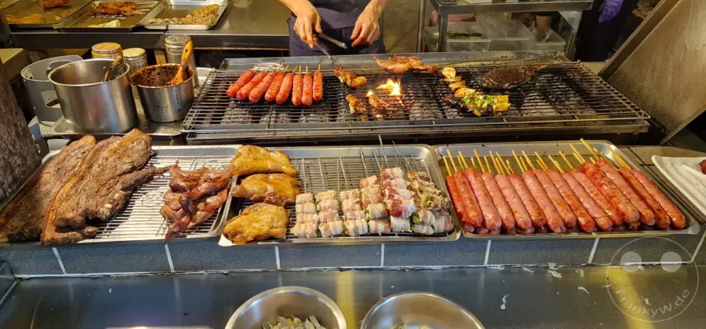 In Jiufen Old Street, Taiwan, brutzeln auf einem Grill verschiedene Grillspezialitäten wie Würste und Fleischspieße. Die Szene vermittelt die lebhafte Atmosphäre eines traditionellen taiwanischen Streetfood-Marktes, der mit authentischen und herzhaften Gerichten lockt.