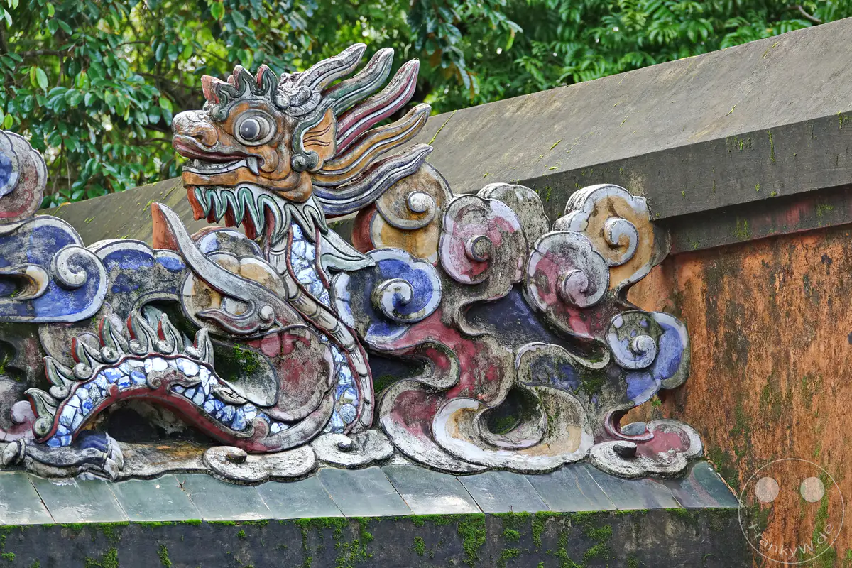Eine detailreiche und farbenfrohe Drachenskulptur ziert die Dachlinie des Grabmals von Tu Duc in Hue, Vietnam. Die kunstvolle Verzierung mit stilisierten Wolken und traditionellen Motiven spiegelt die reiche kulturelle Geschichte des Ortes wider. Moosbewuchs und verwitterte Oberflächen verleihen der Szene eine authentische und atmosphärische Note vergangener Zeiten.