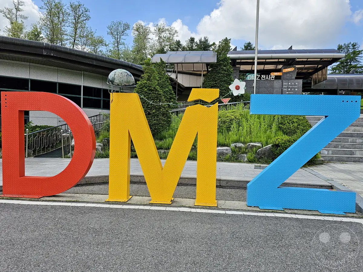 Die Demilitarisierte Zone (DMZ) zwischen Nord- und Südkorea wird hier durch eine auffällige Buchstabeninstallation symbolisiert. Im Hintergrund erhebt sich das DMZ-Pavillon-Gebäude, umgeben von üppiger Vegetation und einem blauen Himmel mit Wolken. Die Szene vermittelt ein Gefühl der Geschichte und des Friedens, trotz der angespannten politischen Lage der Region.