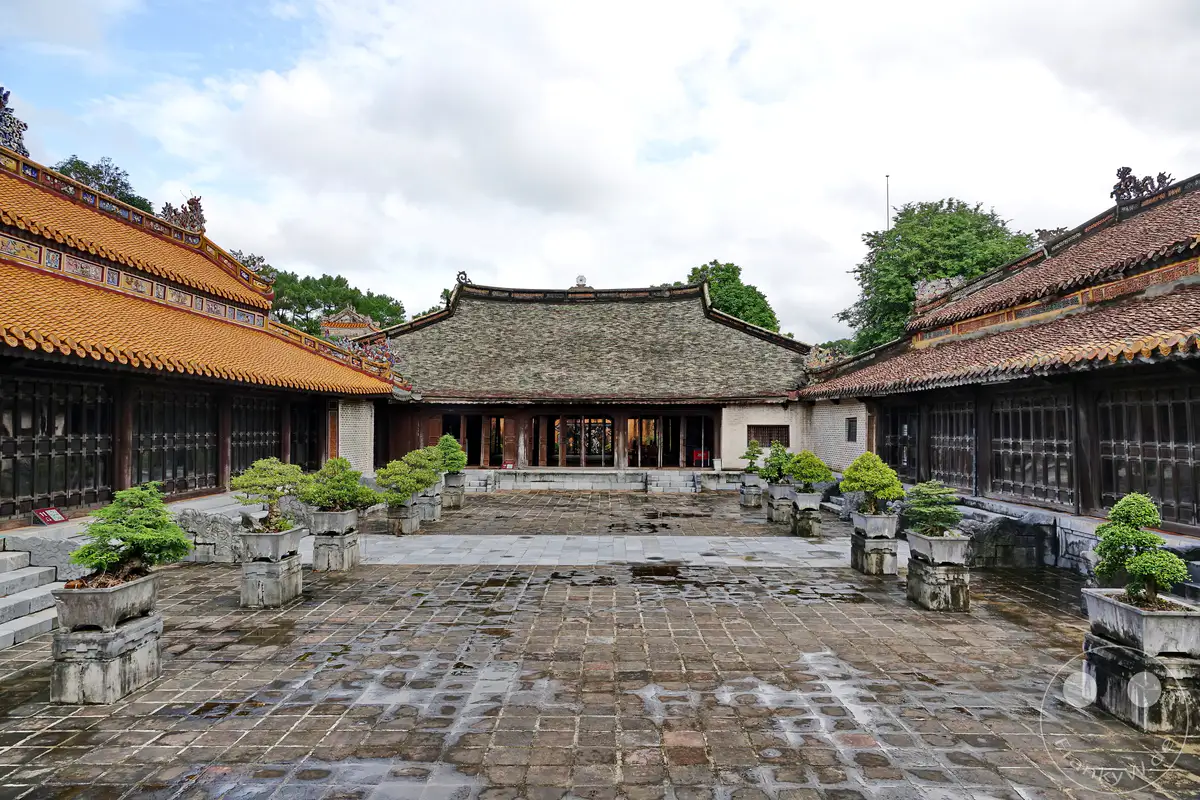 Vietnam - Hue zeigt den Courtyard des Grabmals von Tu Duc, eine Stätte kaiserlicher Architektur. Die Szene strahlt eine ruhige und historische Atmosphäre aus, verstärkt durch die nassen Pflastersteine und die sorgfältig arrangierten Bonsai-Bäume. Grüne Bäume und eine bewölkte Himmelskulisse runden das Bild ab und unterstreichen die Erhabenheit des Ortes.