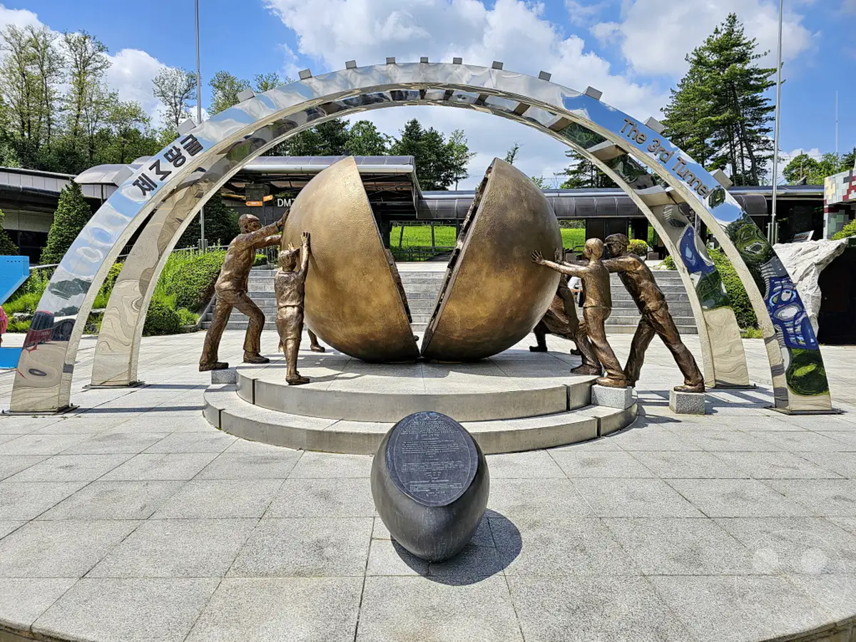 In Südkorea, nahe der DMZ, erinnert eine Bronzeskulptur an den Dritten Tunnel. Mehrere Figuren schieben eine riesige, halbrunde Kugel, die einen Tunnel symbolisiert. Die polierte Metallkonstruktion im Hintergrund reflektiert die Umgebung und verstärkt die feierliche Atmosphäre dieses Ortes.