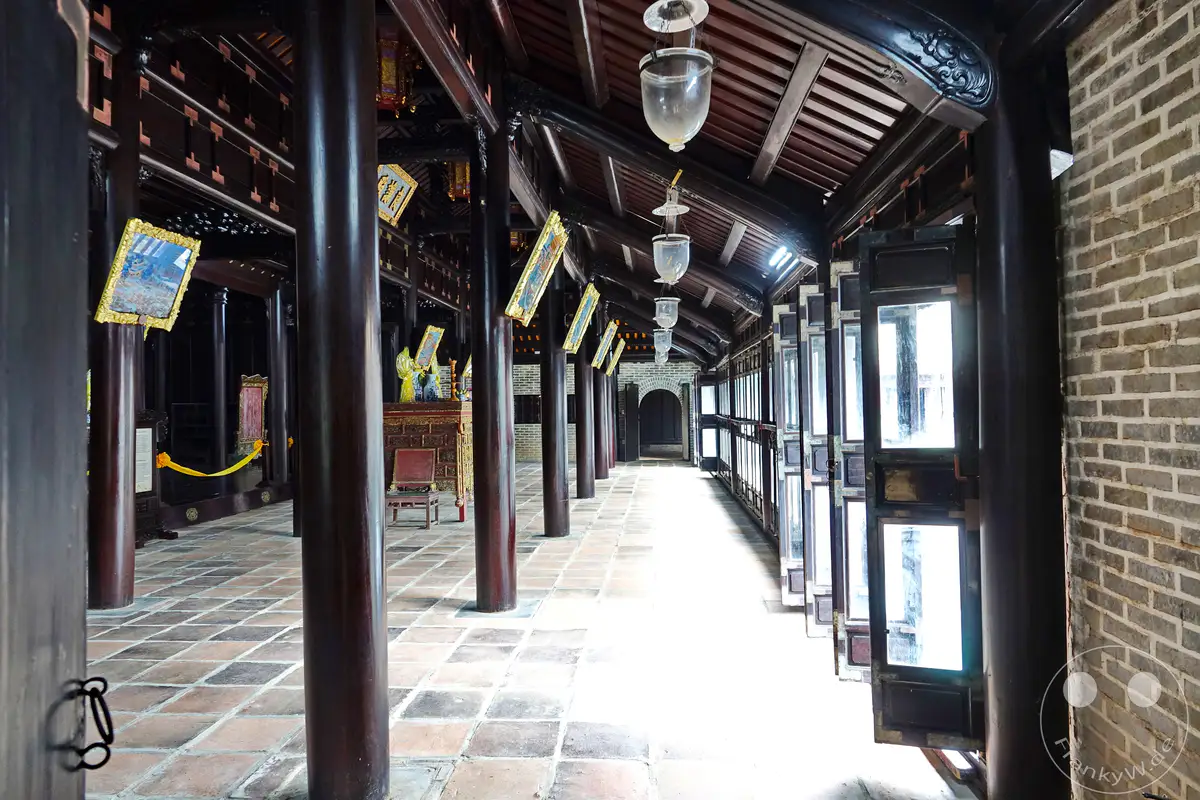 Das Grabmal von Tu Duc in Hue, Vietnam, offenbart eine harmonische Verbindung aus traditioneller Architektur und einer beruhigenden Atmosphäre. Der lange Gang mit seinen zahlreichen Türen und Fenstern, gesäumt von dunklen Holzpfeilern und einer Ziegelwand, führt zu einem Bereich, der von Gemälden und antiken Artefakten geschmückt ist. Die Szene strahlt eine tiefe historische Bedeutung und eine Aura der Ruhe aus, die die Pracht vergangener Zeiten widerspiegelt.