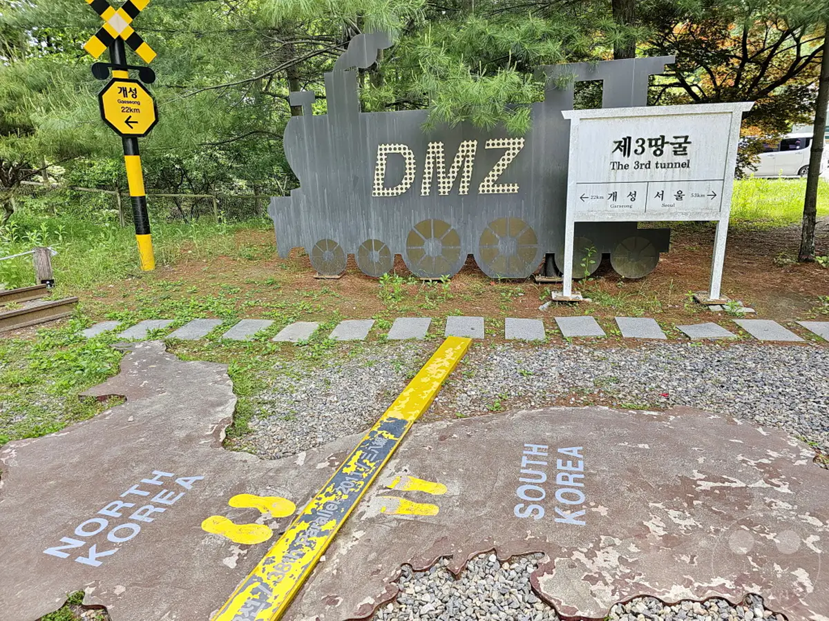 Ein Kunstwerk im Stil einer Dampflokomotive mit der Aufschrift "DMZ" steht vor einem Schild, das auf den 3. Tunnel hinweist. Auf dem Boden ist eine stilisierte Karte der koreanischen Halbinsel mit den Bezeichnungen "North Korea" und "South Korea" dargestellt. Eine gelbe Markierung und Schienen deuten auf die historische Bedeutung des Ortes hin.