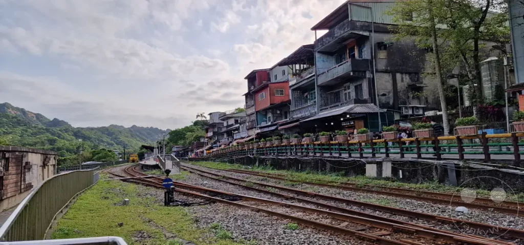 Bahngleise führen an einer Reihe von bunten Häusern vorbei, die sich an einem Hang in Taiwan schmiegen. Die Gebäude sind eng aneinandergereiht und vermitteln ein lebendiges, aber auch etwas chaotisches Bild. Im Hintergrund erheben sich grüne Berge unter einem bewölkten Himmel, was der Szene eine melancholische Note verleiht.