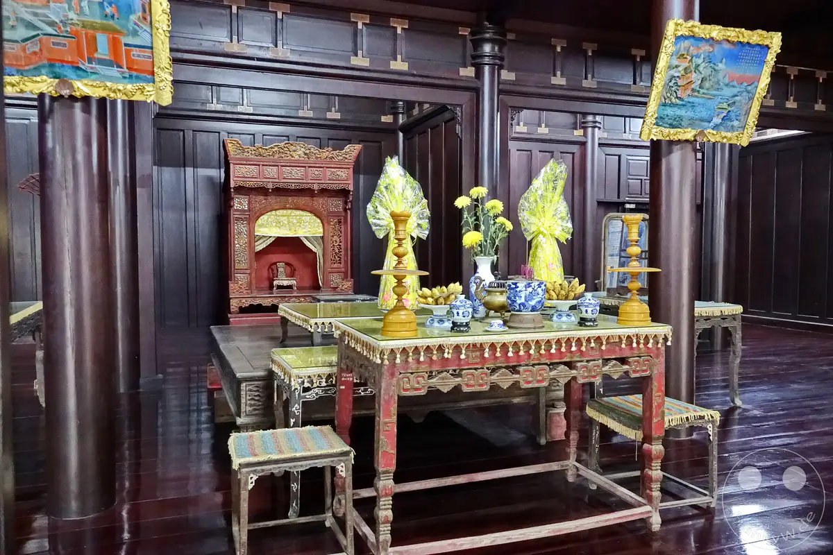 In der kaiserlichen Residenz in Hue, Vietnam, wird ein prunkvoller Altar mit Opfergaben und Gebetsutensilien präsentiert. Die Szene vermittelt eine feierliche und historische Atmosphäre, die tief in der vietnamesischen Kultur verwurzelt ist. Dunkles Holz und aufwendige Schnitzereien rahmen den Zeremonialbereich ein, der an die Pracht des Tu Duc Grabmals erinnert.