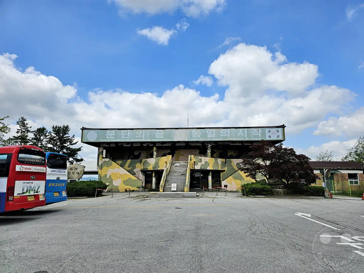 Ein Gebäude mit Tarnmuster und koreanischer Schrift trägt die Aufschrift "End of Separation, Beginning of Unification". Vor dem Gebäude stehen zwei Reisebusse, einer davon deutlich mit "DMZ" beschriftet. Die Szene vermittelt eine Mischung aus touristischer Aktivität und der historischen Bedeutung des Ortes an der Grenze zwischen Nord- und Südkorea in Paju.