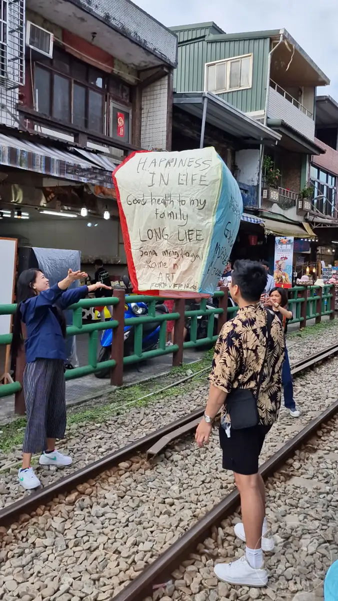 In Taiwan, nahe der Jiufen Old Street, werden Himmelslaternen mit persönlichen Wünschen beschriftet und zum Himmel geschickt. Ein junges Paar beteiligt sich an diesem Ritual, während im Hintergrund belebte Gassen und traditionelle Gebäude zu sehen sind. Die Szene strahlt eine Mischung aus Hoffnung und kultureller Verbundenheit aus, festgehalten an diesem besonderen Ort.
