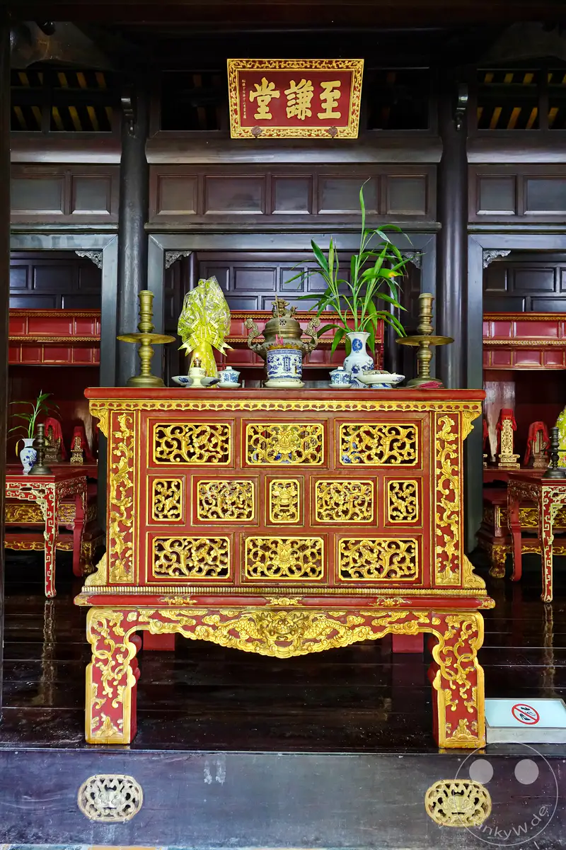 Im Inneren des Grabmals von Tu Duc in Hue, Vietnam, steht eine prächtige, rot und gold verzierte Truhe im Zentrum eines traditionellen Tempelraums. Auf der Truhe sind verschiedene Kultgegenstände wie eine Weihrauchurne, Vasen und Kerzenständer arrangiert, was eine Atmosphäre der Andacht und historischen Ehrfurcht schafft. Die aufwendigen Holzschnitzereien und die dunkle, glänzende Holzvertäfelung spiegeln die kaiserliche Ästhetik und die reiche Kultur Vietnams wider.
