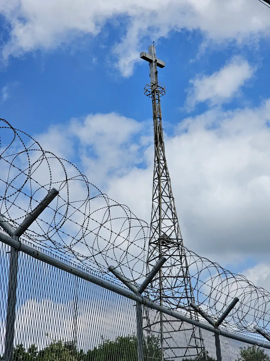 Ein imposanter Kirchturm mit einem Kreuz ragt vor einem dramatischen Himmel mit Wolken in die Höhe. Stacheldraht und ein Gitterzaun trennen den Vordergrund ab und symbolisieren die angespannte Grenze. Die Szenerie vermittelt ein Gefühl von Isolation und spiritueller Wachsamkeit in der entmilitarisierten Zone Südkoreas.