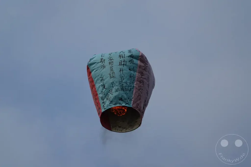 Eine bunte Himmelslaterne mit chinesischen Schriftzeichen steigt in den Nachthimmel über Taiwan auf. Die leuchtende Flamme am unteren Ende der Laterne sorgt für eine warme und feierliche Atmosphäre. Dieses traditionelle Schauspiel ist ein typisches Ereignis in der Nähe der Jiufen Old Street.