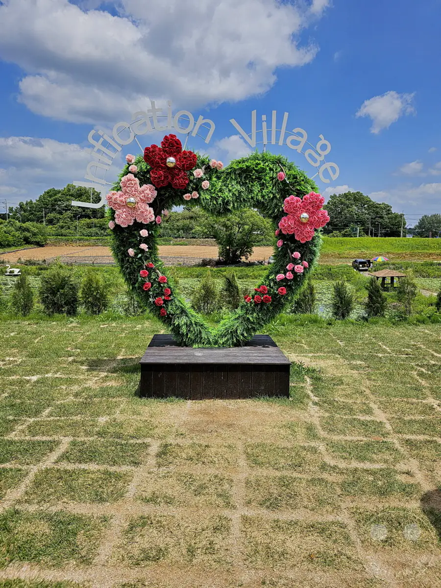 Eine imposante Herzskulptur, geschmückt mit roten und rosa Blumen, steht im Vordergrund eines ländlichen Parks in Paju, Südkorea. Hinter der Dekoration sind üppige grüne Bäume und eine weite Himmelslandschaft mit Wolken zu sehen, was eine friedliche und romantische Atmosphäre schafft. Der Parkbereich ist mit Gras bedeckt und von gepflasterten Wegen durchzogen, die die Szenerie ergänzen.