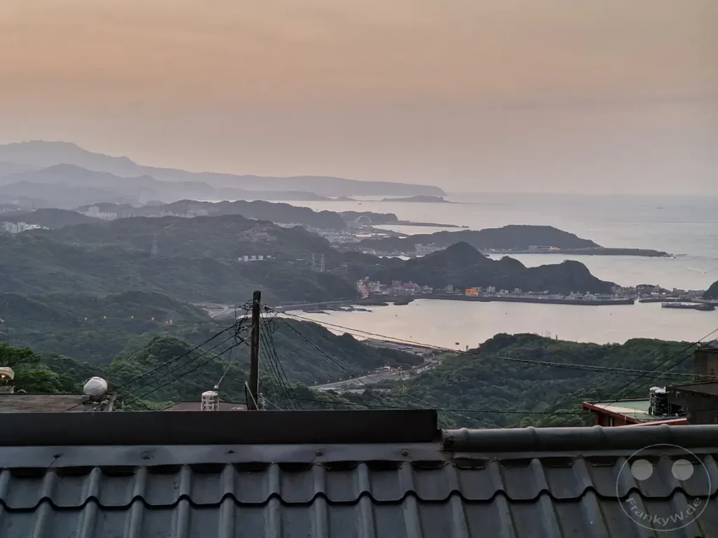 Eine weite Küstenlandschaft offenbart sich in den Dämmerungsstunden, während die sanften Hügel sich bis zum ruhigen Meer erstrecken. Lichter der Stadt Jiufen und der umliegenden Dörfer spiegeln sich im Wasser und erzeugen eine friedliche Atmosphäre. Die dramatische Silhouette der Berge im Hintergrund bildet einen stimmungsvollen Abschluss dieses malerischen Ausblicks in Taiwan.