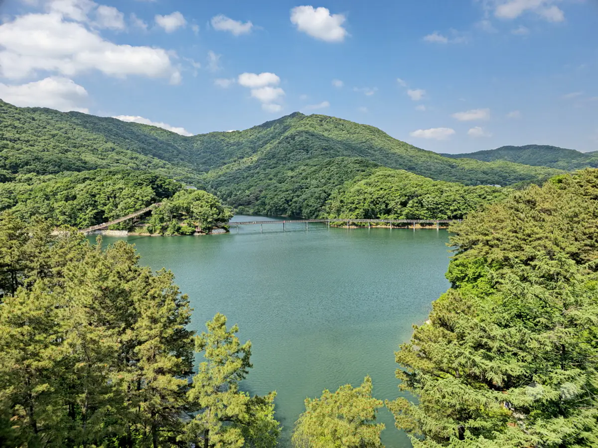 Ein ruhiger See, umgeben von üppigen grünen Bergen, bietet eine friedliche Kulisse in Südkorea. Eine Fußgängerbrücke erstreckt sich über das Wasser und lädt zu einem Spaziergang durch diese malerische Landschaft ein. Die Szene strahlt eine tiefe Ruhe und natürliche Schönheit aus.