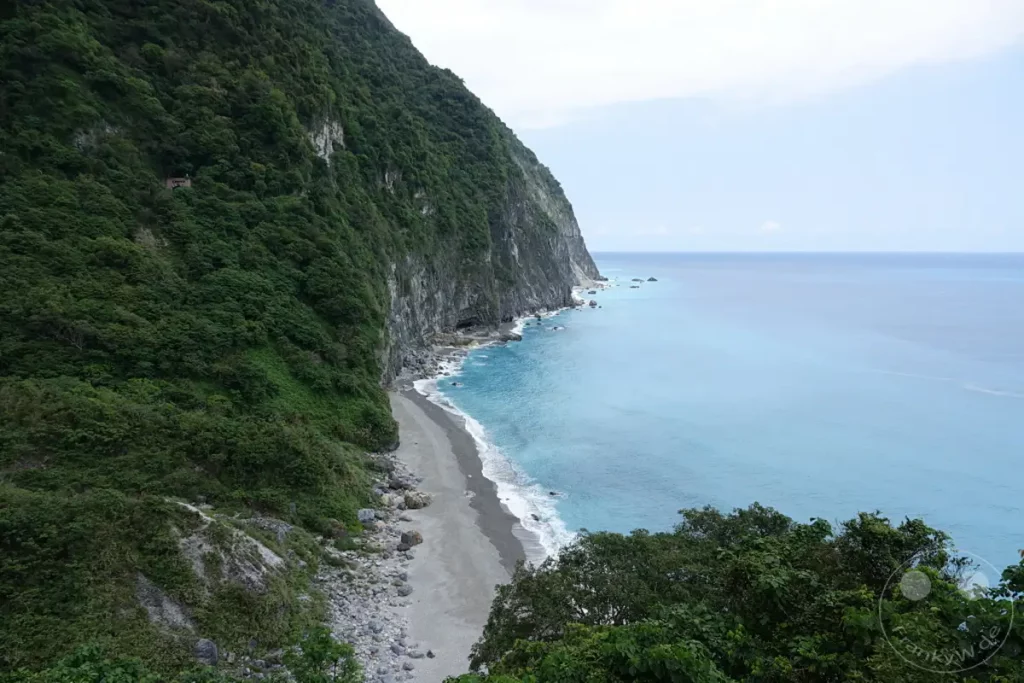 Die majestätische Steilküste von Qingshui Cliff in Taiwan erhebt sich dramatisch über dem türkisfarbenen Pazifik. Wellen brechen sanft an einem Kiesstrand, der sich entlang des Fuße der üppig bewachsenen Felswand erstreckt. Die Szenerie strahlt eine beeindruckende Ruhe und natürliche Erhabenheit aus, die zum Staunen einlädt.