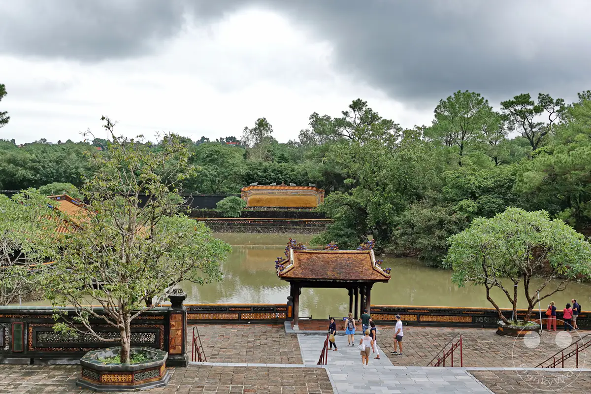 Vietnam - Hue - Das Grabmal von Tu Duc, eine bedeutende historische Stätte, präsentiert sich inmitten üppiger Vegetation und einem ruhigen Gewässer. Touristen erkunden die traditionelle Architektur mit ihren geschwungenen Dächern und verzierten Mauern, die eine Atmosphäre von Gelassenheit und vergangener Pracht vermitteln. Die Szene wird von einem bedeckten Himmel unterstrichen, der der Landschaft eine melancholische und zugleich friedliche Stimmung verleiht.