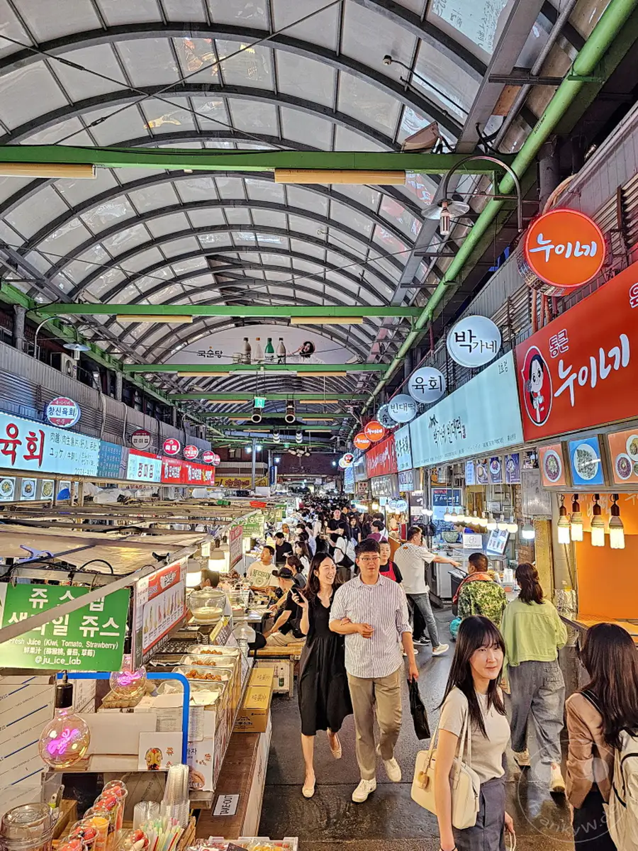 Menschen schlendern durch den belebten Gwangjang Market in Seoul, Südkorea, flankiert von zahlreichen Essensständen, die eine Vielzahl von Streetfood anbieten. Die bogenförmige Decke des Marktes und die bunten Leuchtreklamen schaffen eine lebhafte und einladende Atmosphäre. Kulinarische Genüsse und die geschäftige Stimmung laden Besucher ein, die lokale Esskultur zu erleben.