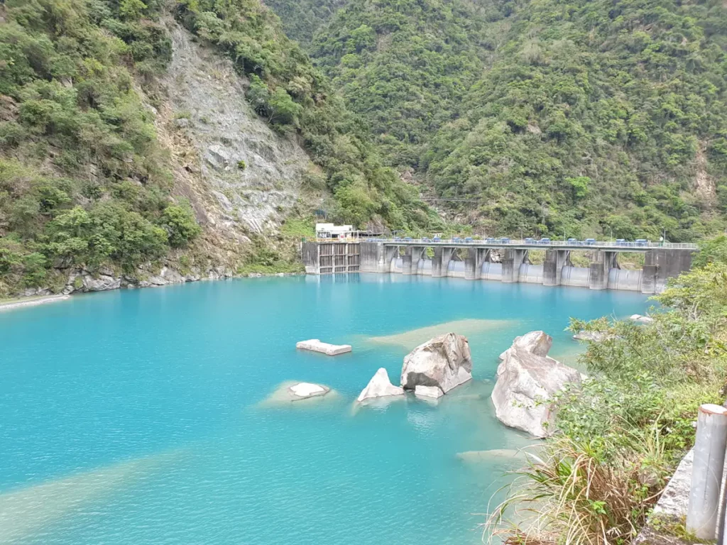 Türkisfarbenes Wasser fließt um Felsen herum und mündet in einen imposanten Staudamm, der sich unter einer üppig grünen Bergkulisse erstreckt. Die Szenerie in Taiwan, im Herzen des Taroko Nationalparks, strahlt eine friedliche und zugleich kraftvolle Atmosphäre aus. Ein beeindruckendes Zusammenspiel von Natur und Ingenieurskunst.