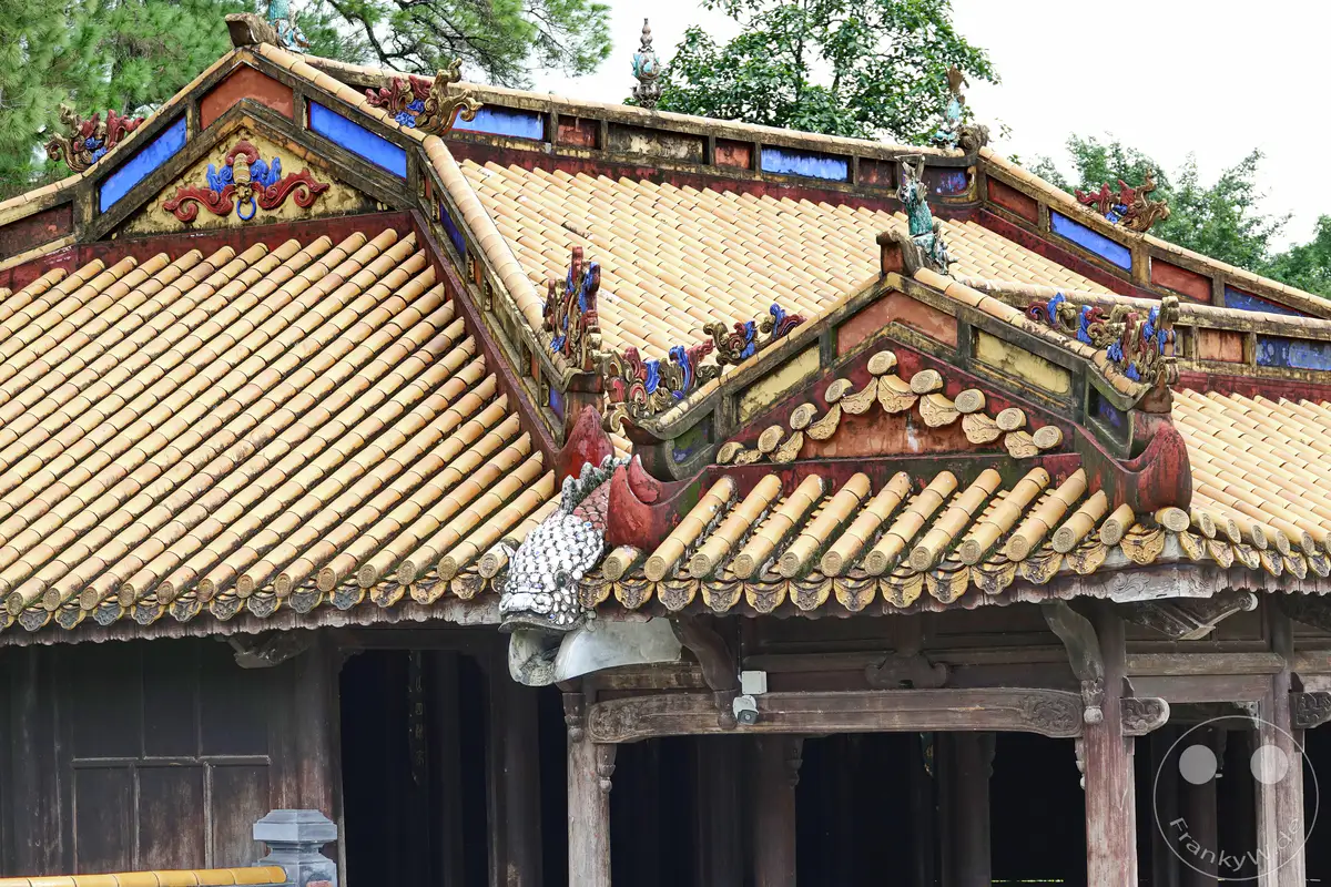 Ein detailreicher Blick auf das reich verzierte Dach des Kaisergrabmals von Tu Duc in Hue, Vietnam. Die traditionelle vietnamesische Architektur mit ihren gelben Ziegeln und bunten Keramikornamenten strahlt historische Bedeutung aus. Die Szene vermittelt eine Atmosphäre von Ehrfurcht und kulturellem Erbe inmitten üppiger Vegetation.