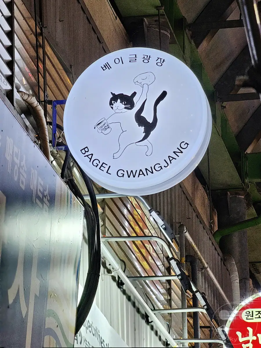 Ein runde beleuchtete Leuchtreklame mit einer stilisierten Katze, die einen Bagel hält, hängt über dem Gwangjang Market in Seoul, Südkorea. Die Reklame wirbt für "BAGEL GWANGJANG" und trägt koreanische Schriftzeichen am oberen Rand. Die nächtliche Szene unterstreicht den Charme des geschäftigen Marktes mit seinen vielfältigen kulinarischen Angeboten.