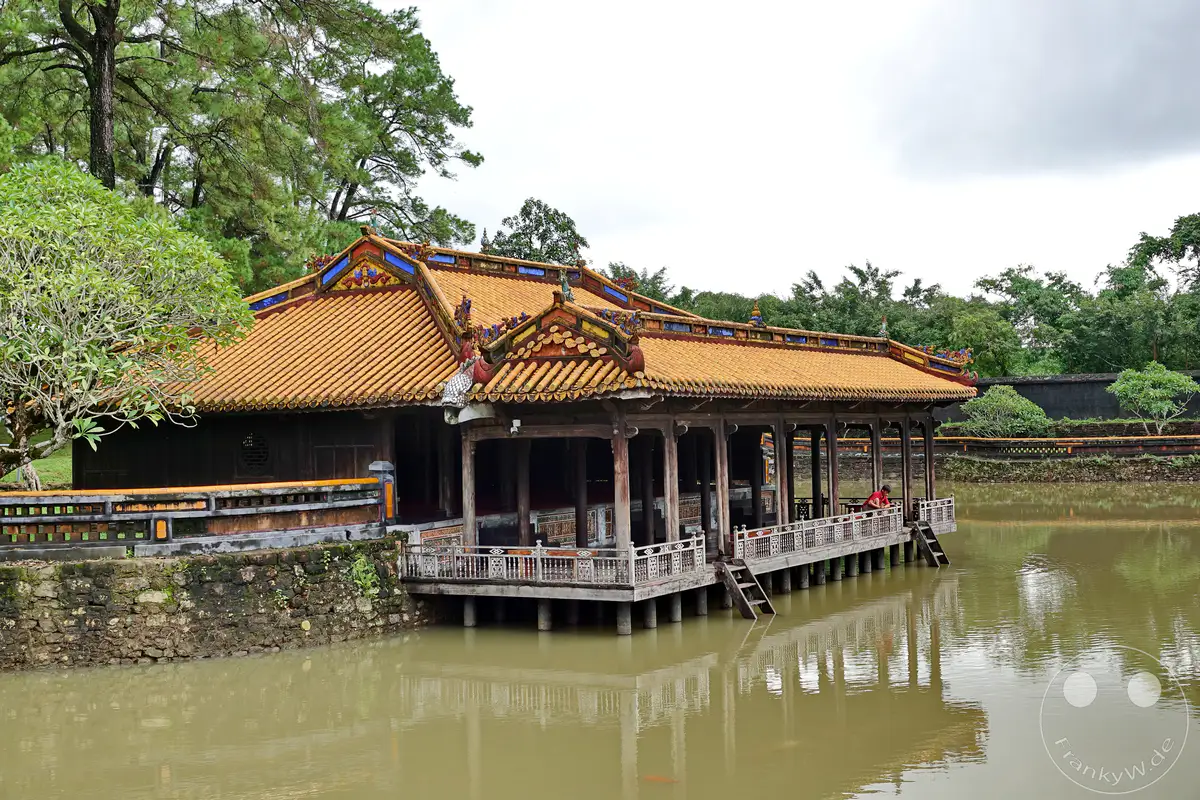 In Hue, Vietnam, erhebt sich eine traditionelle Pagodenstruktur mit charakteristischen ockerfarbenen Ziegeln über einem ruhigen Teich. Die auf Stelzen erbaute überdachte Veranda mit filigranen Holzschnitzereien bietet einen Einblick in die kaiserliche Architektur und strahlt eine friedliche, historische Atmosphäre aus. Die Spiegelungen der üppigen Vegetation und des Gebäudes auf der Wasseroberfläche verstärken die malerische Szenerie.