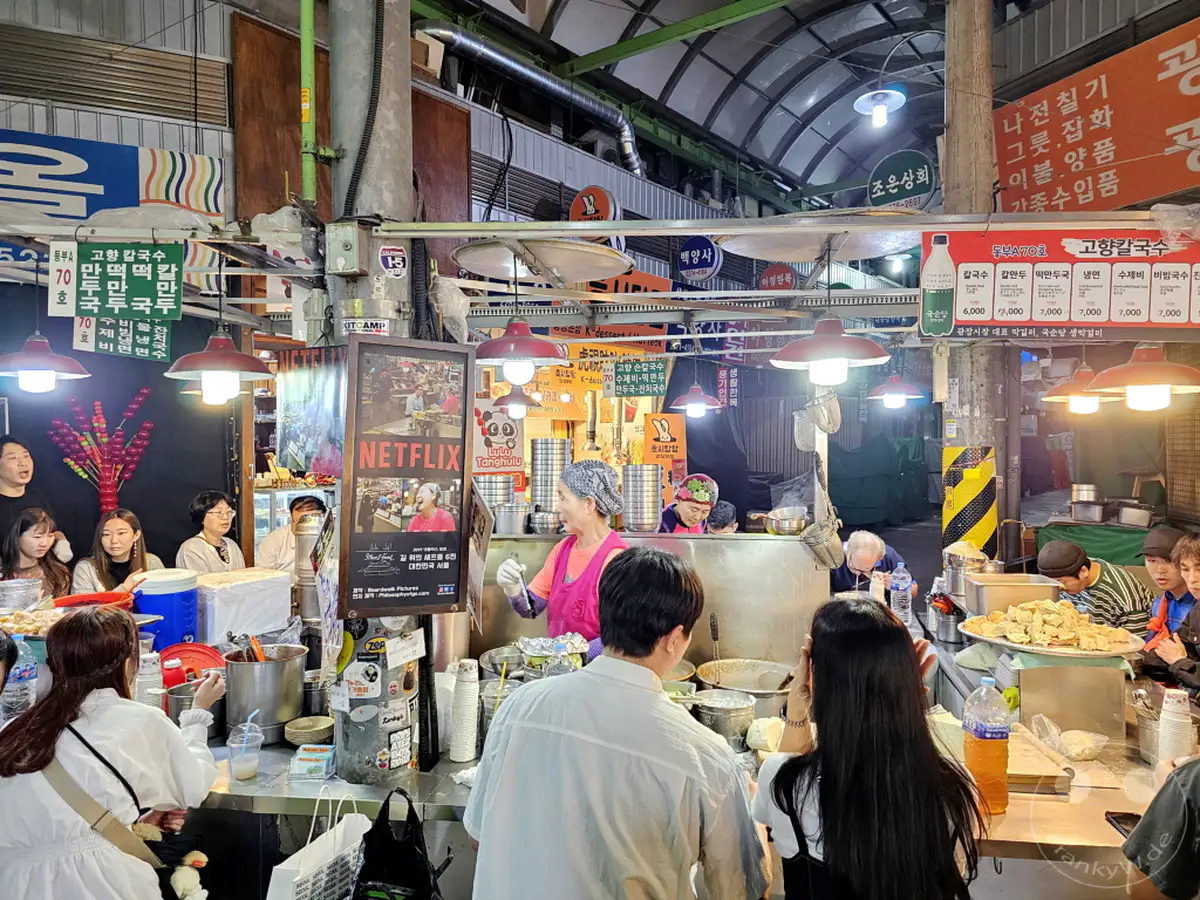 Der Gwangjang Market in Seoul, Südkorea, pulsiert vor Leben mit zahlreichen Ständen, die eine Vielfalt an authentischem koreanischem Street Food anbieten. Menschen drängen sich an den Verkaufsflächen, um herzhafte und süße Leckereien zu probieren, was eine geschäftige und lebendige Marktatmosphäre schafft. Das bunte Treiben und die dampfenden Speisen laden zum kulinarischen Entdecken ein.