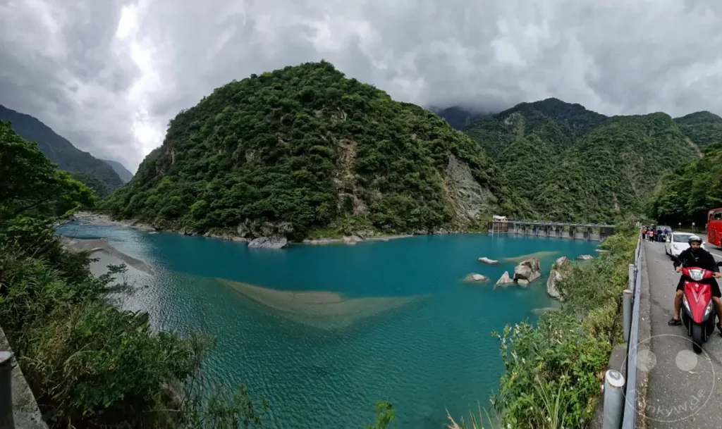 Türkisfarbene Flusswasser schlängeln sich durch die majestätische Schlucht des Taroko Nationalparks in Taiwan, flankiert von üppig grünen Bergen und überragt von einer dramatischen Wolkendecke. Ein imposanter Damm markiert die Kulisse, während auf der Straße am Ufer Verkehr herrscht, darunter ein Motorradfahrer und ein Bus. Die Szenerie strahlt eine beeindruckende und doch leicht bedrohliche Naturgewalt aus.
