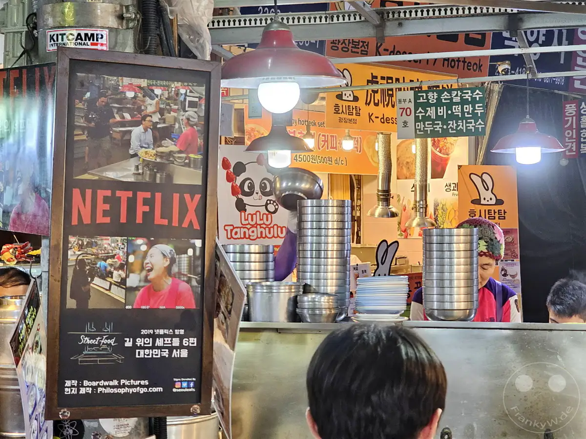 Eine leuchtende Netflix-Werbetafel im geschäftigen Gwangjang Market in Seoul, Südkorea, lockt mit Bildern von Street Food und ikonischen Netflix-Szenen. Im Hintergrund sind Stände mit verschiedenen koreanischen Spezialitäten und eine freundliche Verkäuferin zu sehen, die ein Lächeln zeigt. Die Atmosphäre ist lebhaft und lädt dazu ein, die kulinarischen Entdeckungen und die bunte Marktatmosphäre zu genießen.