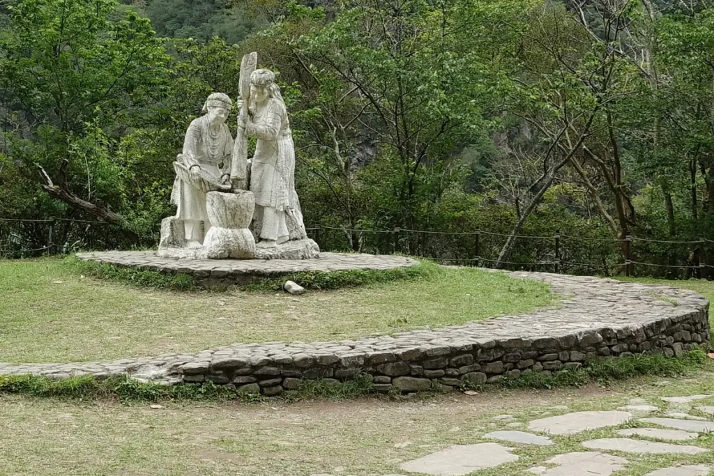 Im Taroko Nationalpark von Taiwan, genauer gesagt in Buluowan, stehen zwei detailgetreue Steinfiguren, die eine Szene traditioneller Handwerkskunst darstellen. Die Figuren und die umgebende Grünfläche sind von üppigen Bäumen und einer bergigen Landschaft eingerahmt, was eine friedliche und naturverbundene Atmosphäre schafft.