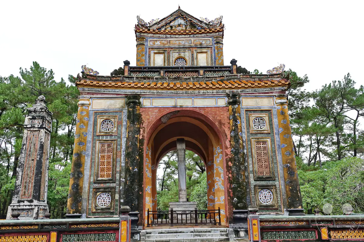 Ein prachtvolles Tor markiert den Eingang zum Grabmal von Tu Duc in Hue, Vietnam. Die detailreiche, bunte Architektur zeugt von kaiserlicher Pracht und kultureller Bedeutung. Die ruhige Atmosphäre wird durch die umliegenden Bäume und den bewölkten Himmel unterstrichen.