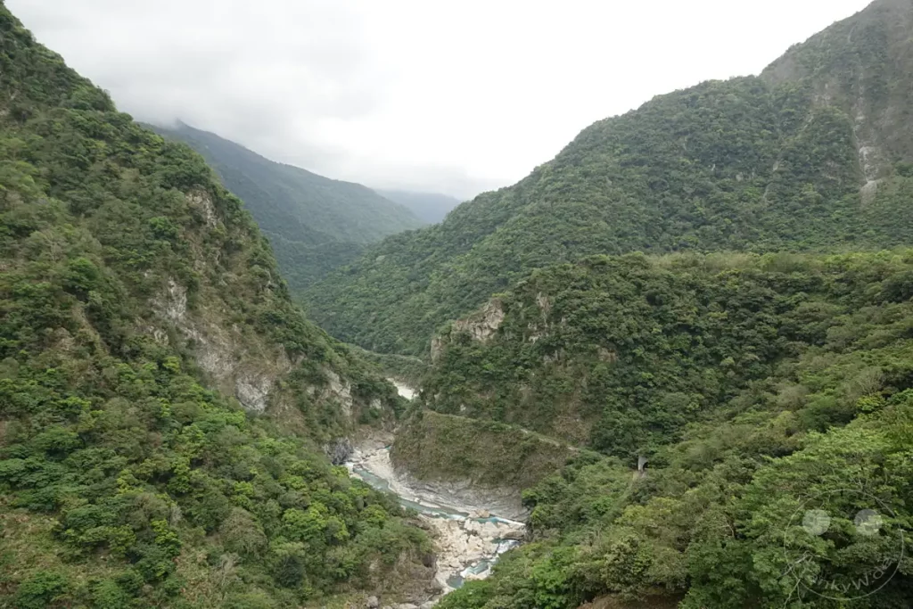 Die majestätische Schlucht des Taroko Nationalparks in Taiwan präsentiert sich in ihrer vollen Pracht, mit steilen, von üppiger Vegetation bedeckten Berghängen, die sich zu beiden Seiten des Taroko-Flusses erheben. Der Fluss schlängelt sich durch das Tal und hinterlässt Spuren von Felsformationen, während die Buluowan Suspension Bridge in der Ferne die Verbindung zwischen den beiden Seiten symbolisiert. Eine friedliche, aber beeindruckende Landschaft, die die Kraft der Natur und die Weite des Nationalparks einfängt.