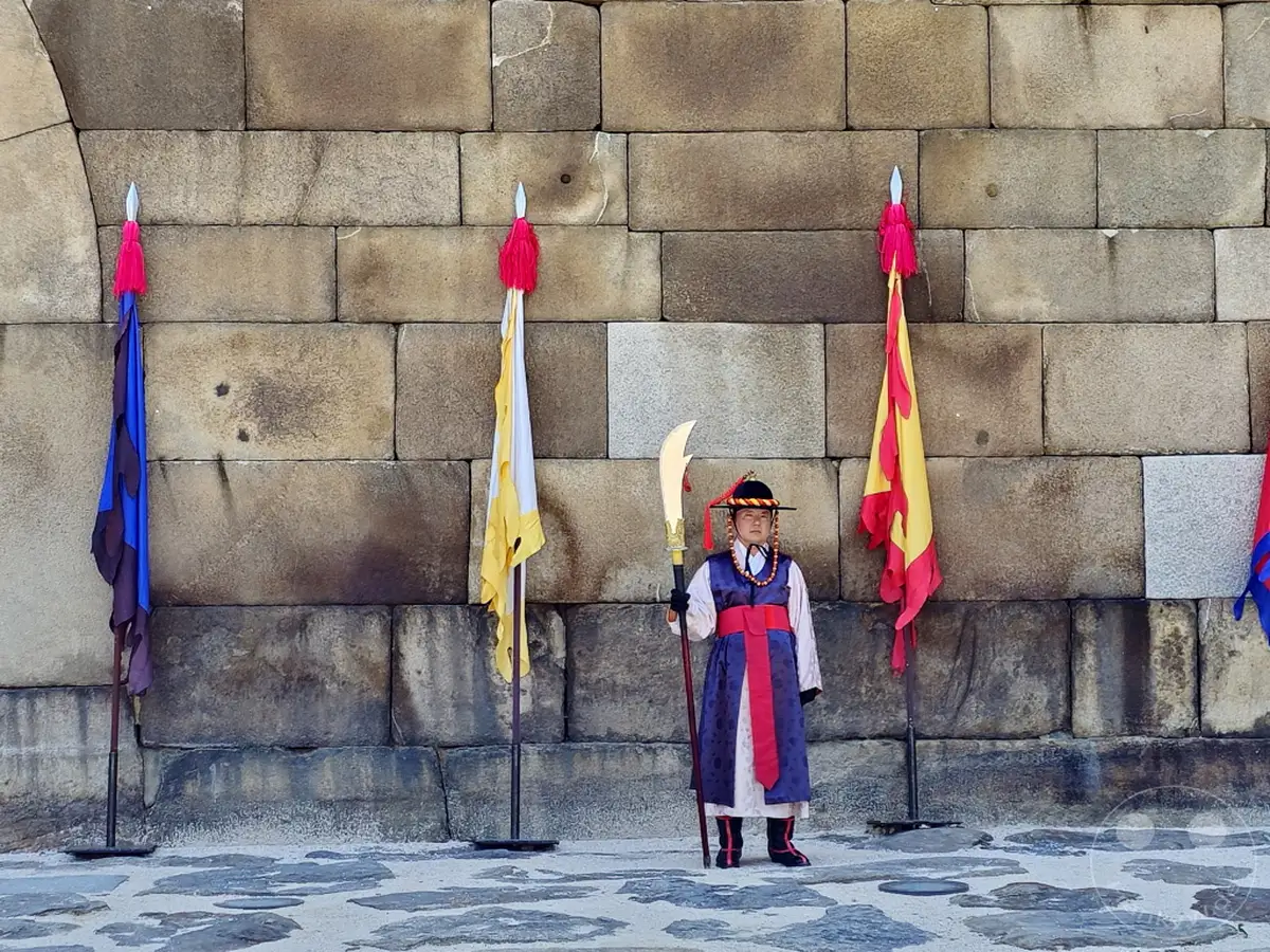 Eine Wachablösungszeremonie findet vor dem Sungnyemun Gate in Seoul statt. Ein Wache in traditioneller Kleidung und Hut steht mit einer zeremoniellen Waffe zwischen bunten Fahnen auf einem Steinpflaster. Die Szene wird durch die imposante Steinmauer im Hintergrund unterstrichen und vermittelt ein Gefühl von Geschichte und kulturellem Erbe.