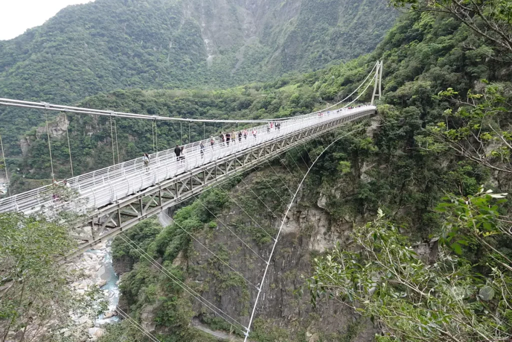 Die Buluowan-Hängebrücke überspannt eine tiefe Schlucht im Taroko-Nationalpark in Taiwan. Touristen spazieren über die breite Brücke und genießen die atemberaubende Aussicht auf die bewaldeten Berghänge und den darunter liegenden Fluss. Die Szenerie strahlt eine friedliche Atmosphäre inmitten dramatischer Naturlandschaften aus.