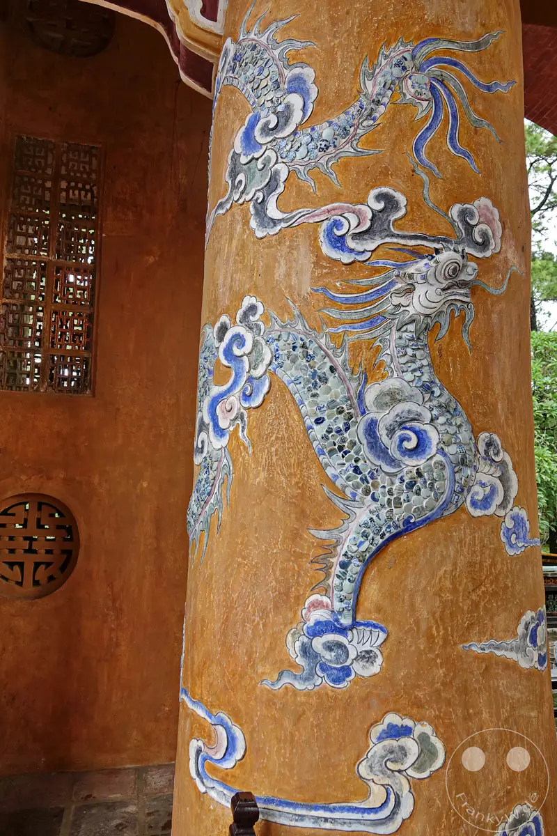 Eine detaillierte Nahaufnahme einer Säule im Grabmal von Tu Duc in Hue, Vietnam, zeigt eine kunstvolle Darstellung eines Drachen aus blauen und weißen Keramikscherben. Wolkenförmige Verzierungen aus bemalter Stuckmasse umgeben die Drachenfigur und erzeugen eine dynamische und traditionelle Atmosphäre. Die leuchtenden Farben und die aufwendige Handwerkskunst spiegeln die kaiserliche Geschichte und die kulturelle Bedeutung des Ortes wider.