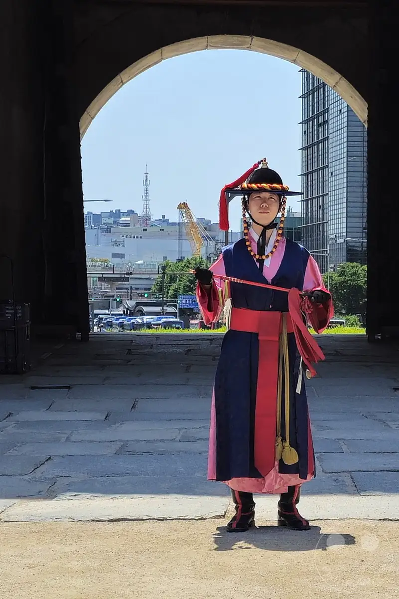 Ein Wachmann in traditioneller koreanischer Kleidung steht vor dem Sungnyemun Gate in Seoul. Die prachtvolle Uniform und die strenge Haltung vermitteln ein Gefühl von Geschichte und Pflichtbewusstsein. Im Hintergrund sind moderne Gebäude und Baukräne zu sehen, die einen interessanten Kontrast zur historischen Kulisse bilden.