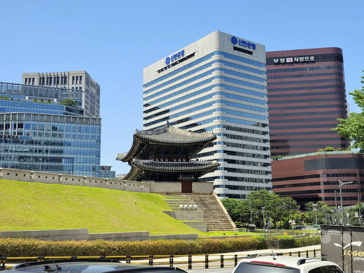 Ein malerischer Blick auf das Sungnyemun Gate in Seoul, Südkorea, vor einer Kulisse moderner Hochhäuser. Die historische Struktur steht im starken Kontrast zu den gläsernen Fassaden der umliegenden Bürogebäude. Die Szene vermittelt eine harmonische Mischung aus Geschichte und Fortschritt unter einem strahlend blauen Himmel.