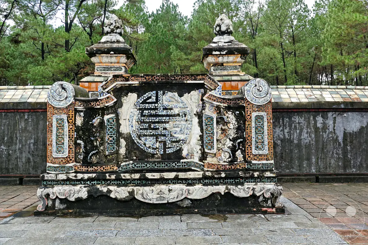 Die aufwendige Architektur des Mausoleums von Tu Duc in Hue, Vietnam, präsentiert sich mit detailreichen Verzierungen und Mosaiken. Die Anlage liegt inmitten einer grünen Waldlandschaft und vermittelt eine Atmosphäre von historischer Bedeutung und erhabener Ruhe. Die verwitterten Materialien und das gedämpfte Licht unterstreichen den zeitlosen Charakter dieser kaiserlichen Ruhestätte.