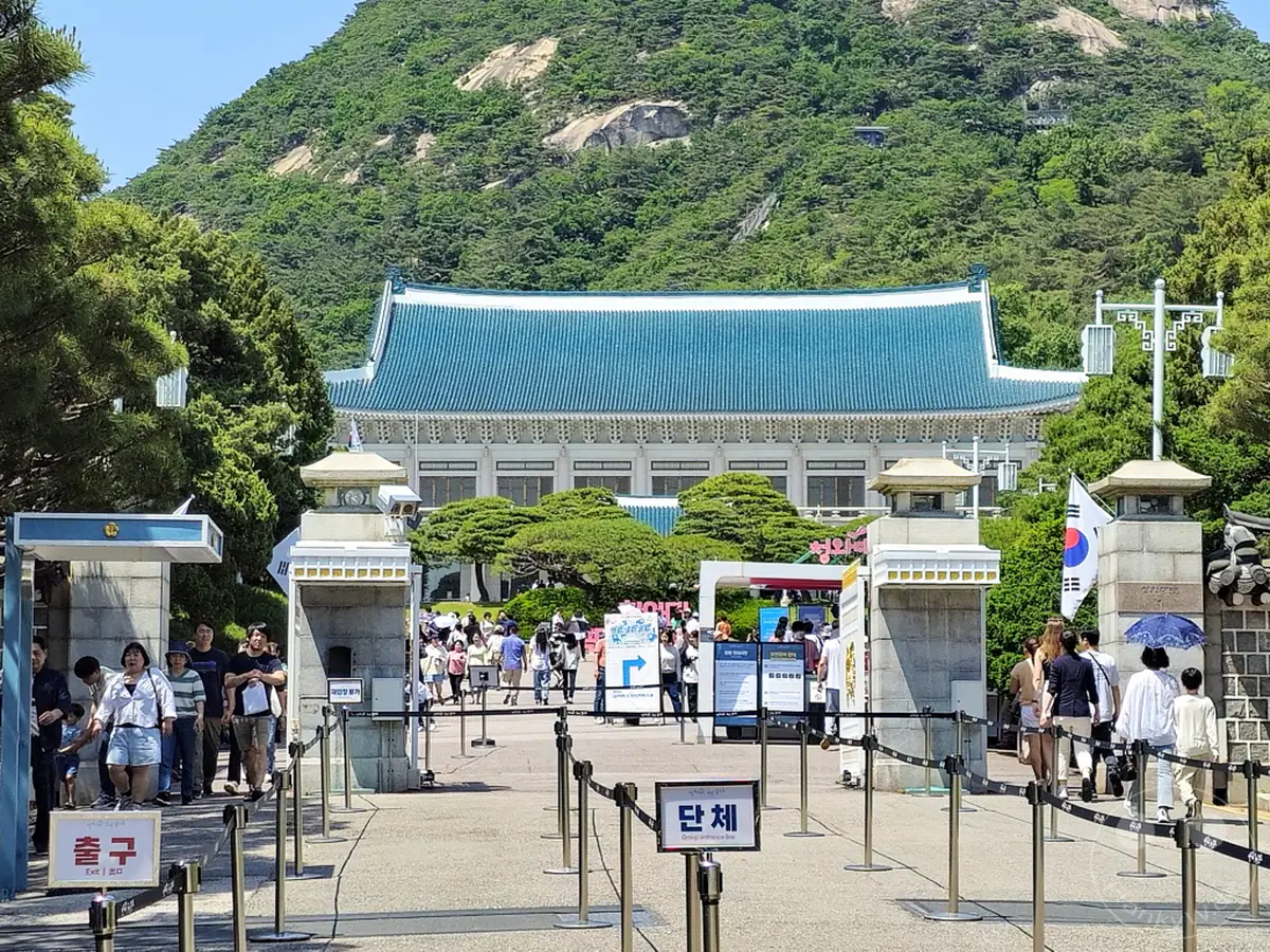 Besucher betreten das Gelände von Cheongwadae, dem ehemaligen Präsidentenpalast in Seoul, Südkorea, auch bekannt als Das Blaue Haus. Menschenmassen bewegen sich durch ein Tor in Richtung des imposanten Gebäudes mit der charakteristischen blauen Dachkonstruktion, umgeben von üppigem Grün und bewacht von steinernen Säulen. Die Atmosphäre ist sonnig und belebt, was auf einen beliebten Ausflugsort für Touristen hindeutet.