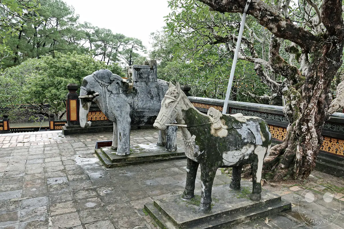 In Hue, Vietnam, befinden sich vor dem Grabmal von Tu Duc zwei alte Steinskulpturen eines Elefanten und eines Pferdes. Die Verwitterung und der Moosbewuchs verleihen den Skulpturen eine geheimnisvolle und ehrwürdige Ausstrahlung. Die gepflasterte Fläche und die umliegende Vegetation verstärken die ruhige und historische Atmosphäre dieses Ortes.