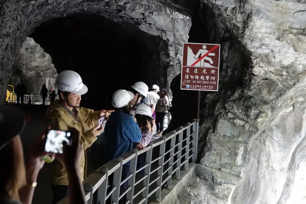 Menschen mit Schutzhelmen stehen auf einem Weg in einem Felsentunnel im Taroko National Park in Taiwan. Ein Warnschild mit der Aufschrift "Cliff! Do Not Cross Over" weist auf die Gefahren am Steilhang hin. Die Szene vermittelt eine Atmosphäre der Vorsicht inmitten beeindruckender Naturformationen.