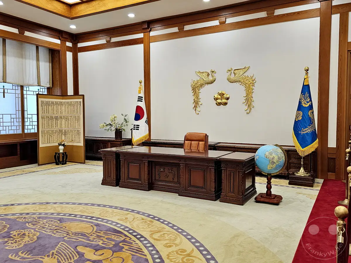 Das imposante Büro von Südkorea im Blauen Haus in Seoul strahlt eine Atmosphäre von Macht und Tradition aus. Ein prunkvoller Schreibtisch mit einem Ledersessel bildet das Zentrum des Raumes, flankiert von den Flaggen Südkoreas und dem Präsidentenamt. Die goldene Dekoration an der Wand und die kunstvollen Teppiche unterstreichen die würdevolle und feierliche Stimmung dieses historischen Ortes.